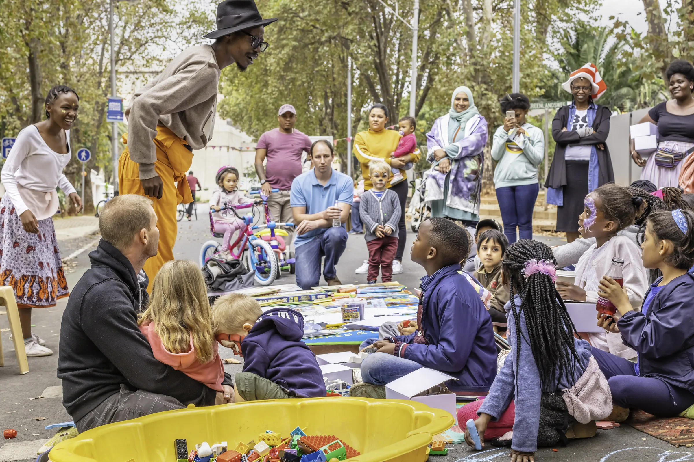 Families and children gathered at an outdoor community event, a man in a hat leaning in to talk to them